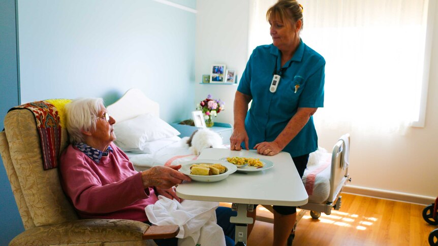 An elderly woman sitting in a chair, with her lunch on a table, talking to a nurse.