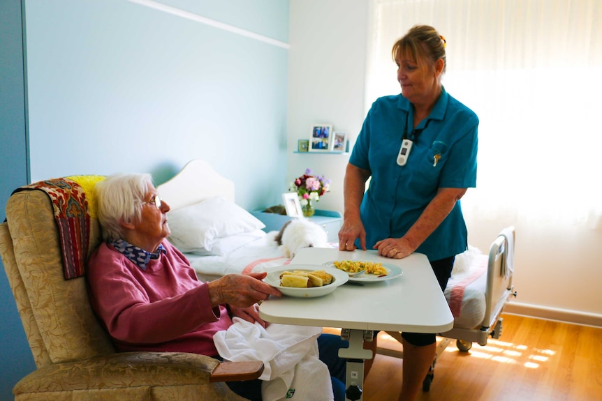 An elderly woman sitting in a chair, with her lunch on a table, talking to a nurse.