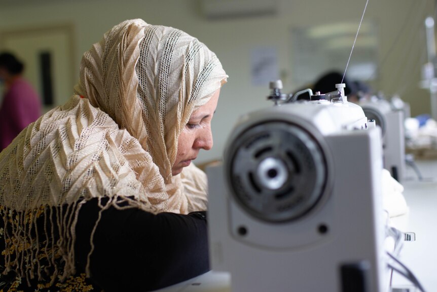 Parima usa un hiyab y trabaja en una máquina de coser blanca cerca de una ventana. 