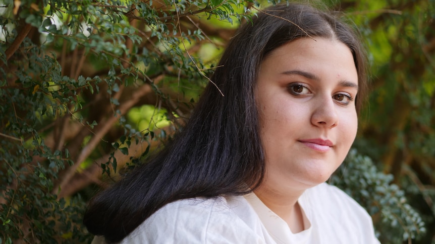 A teenage girl with long brown hair and a white t-shirt looks sideways towwards the camera. Behind her are trees.