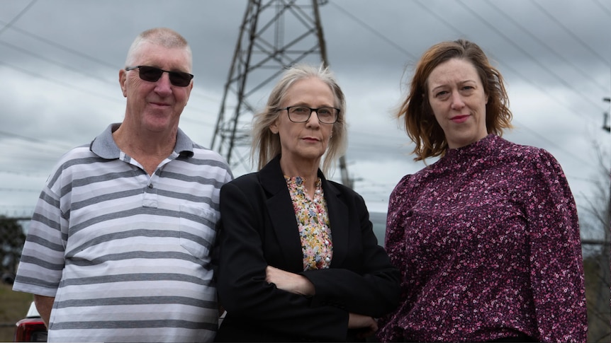 A man and two women stand in front of an electricity pylon