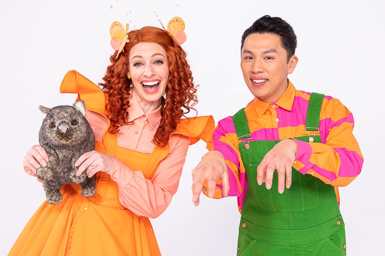 A woman in a bright orange dress holds a wombat as a man in green overalls and an orange and pink shirt makes a wombat sign.