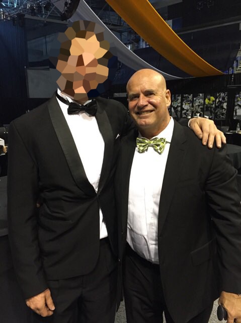 Greg smiles with another person, whose face is pixelated, wearing a bow-tie and suit.