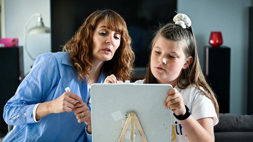 Jorja Mackie (9) writes on a white board as her reading tutor Paula Sciré offers guidance during a lesson at her home, 25 June 2019.