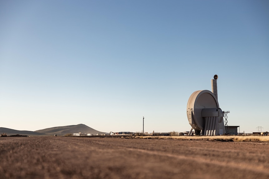 A round space catapult in flat country, light blue sky, hillocks in the background.
