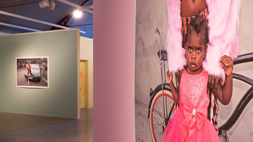 In the foreground a large photo of an Aboriginal toddler wearing a sparkly pink dress hands on a pink gallery wall.