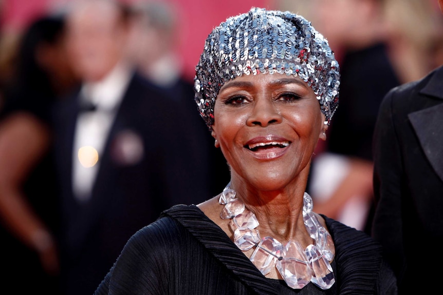 Cicely Tyson smiles as she looks into the camera wearing a sequin hat and what appears to be a resin necklace and a black dress.