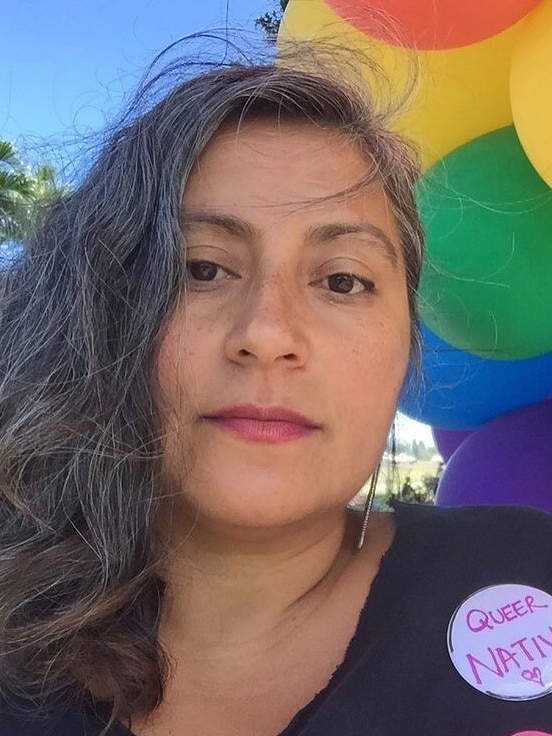 A woman with long grey hair beside an assortment of rainbow coloured balloons on a blue-sky day