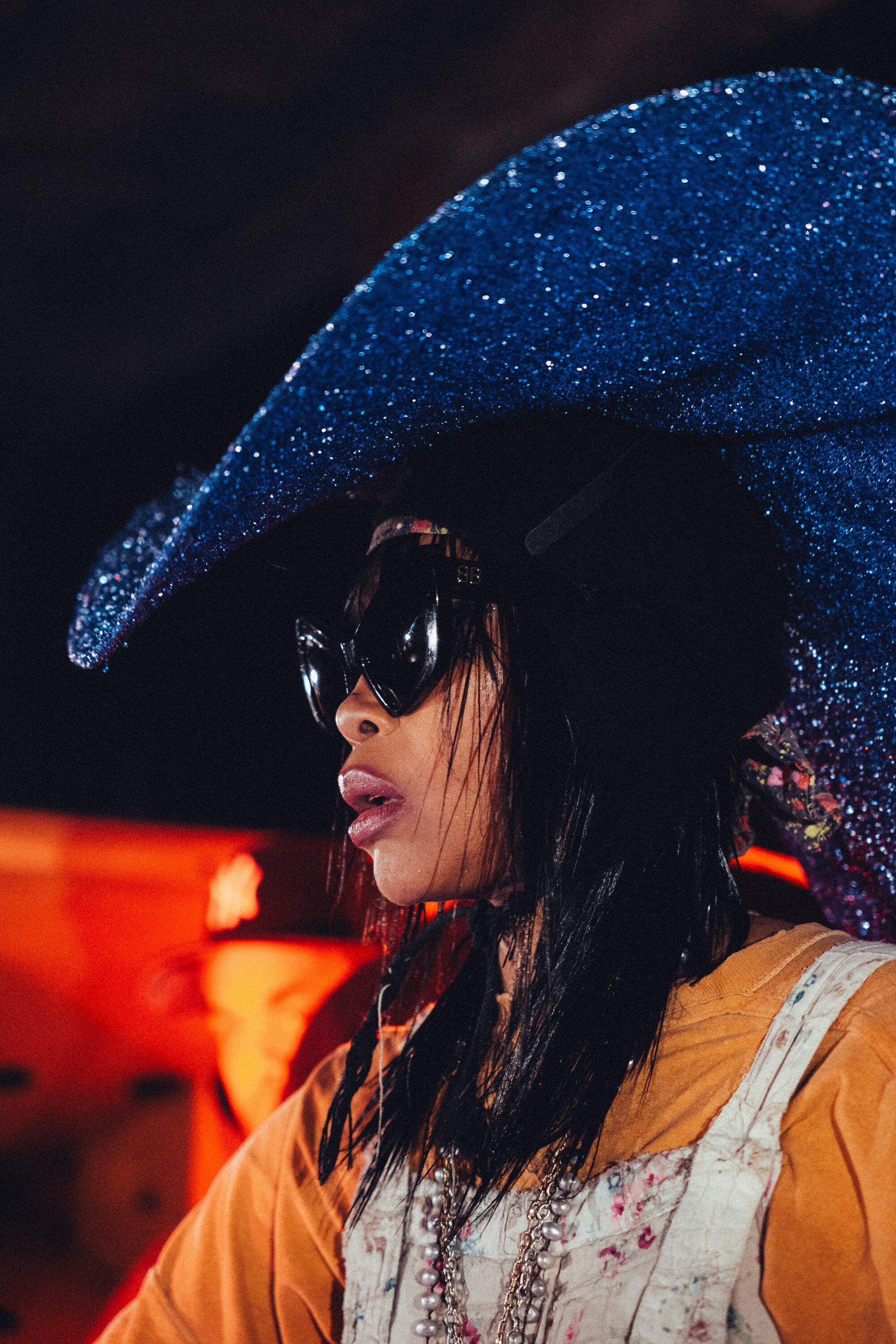 Erykah Badu wearing a large glittery blue hat and layered clothing in warm, dramatic lighting.