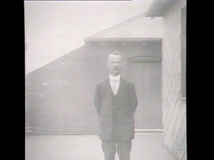 A black and white full body photo of a man in a suit, wearing a tie, against a building.