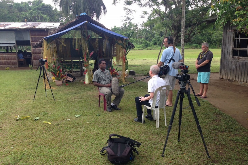 Sean Dorney interviewing school principal in village school with producer and sound recordist watching.