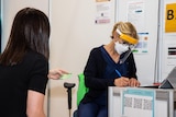 A woman medical professional wearing a mask and PPE makes notes on paper while speaking to a patient