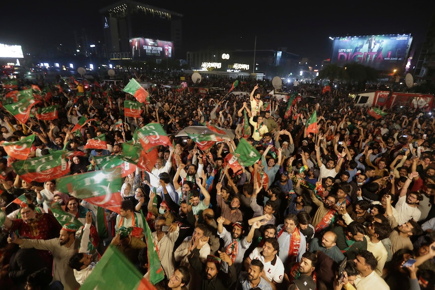 A large crowd of Pakistani people rallying outdoors in the evening, with many wave flags of the Tehreek-e-Insaaf political party