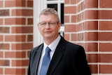 Man with grey hair and glasses dressed in suit stands in front of a red brick building.