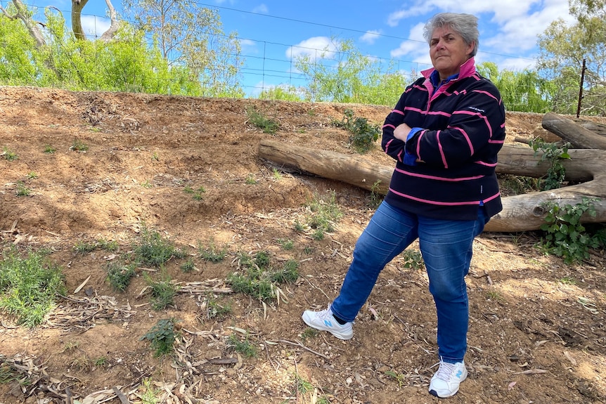Joanne, a woman with short grey hair wearing a pink and navy striped polo shirt stands arms crossed by a levee.