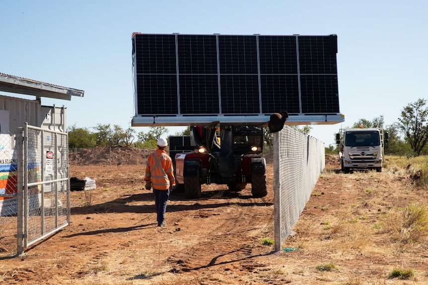 A photo showing a person walking towards a tractor carrying solar panels surround by fence and car outside the fence