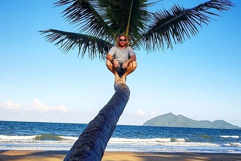 Mr Thomson sits mid-way up a palm tree on the beach with ocean in the background.