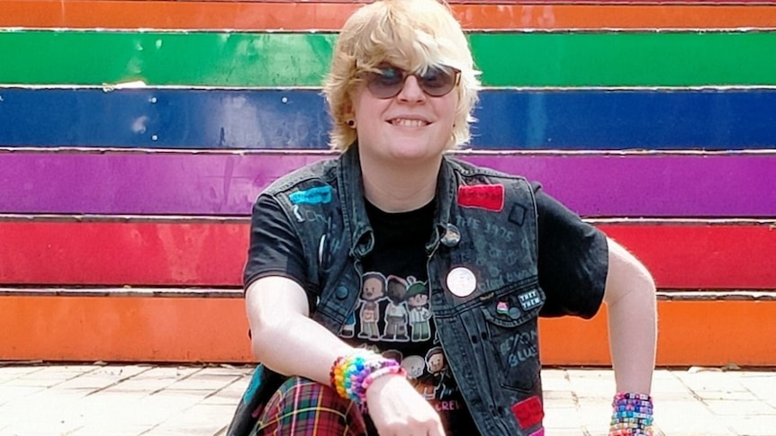A young person sitting on rainbow painted stairs. Short blonde hair, wearing sunglasses