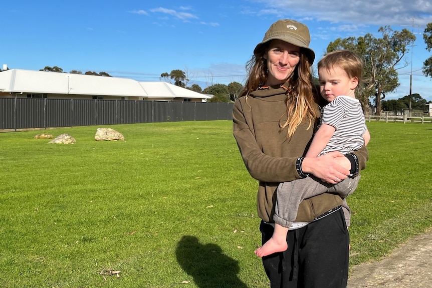 A woman wearing a brown jumper and hat holding a toddler in front of lawn with rocks on it