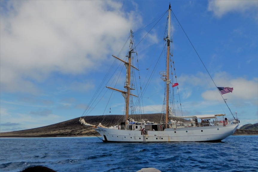 A metal-hulled boat with two masts and a US flag moored next to a low volcanic island