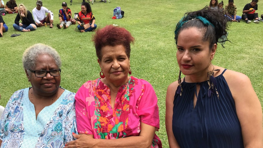 Three Indigenous women sitting in a park, with other people sitting on green grass behind them.