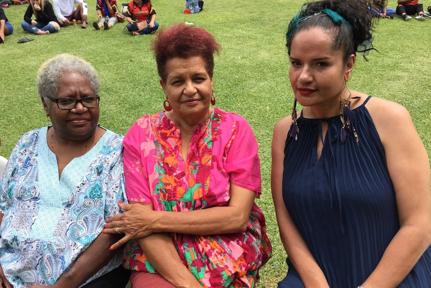 Three Indigenous women sitting in a park, with other people sitting on green grass behind them.