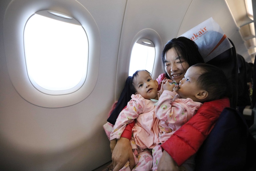 Conjoined twin girsl sit on their mother's lap on an airplane