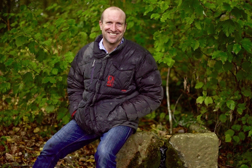 Man with thick jacket on sits on a well in the forest.