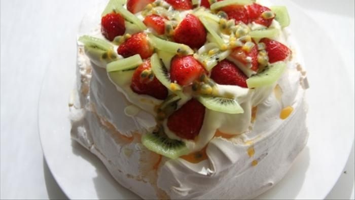 A pavlova covered in whipped cream, kiwi fruit slices and strawberries on a white plate.