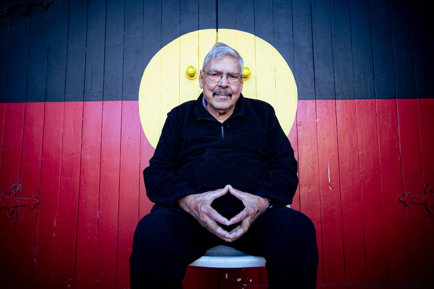 an indigenous man sitting down in front a garage door painted with the aboriginal flag