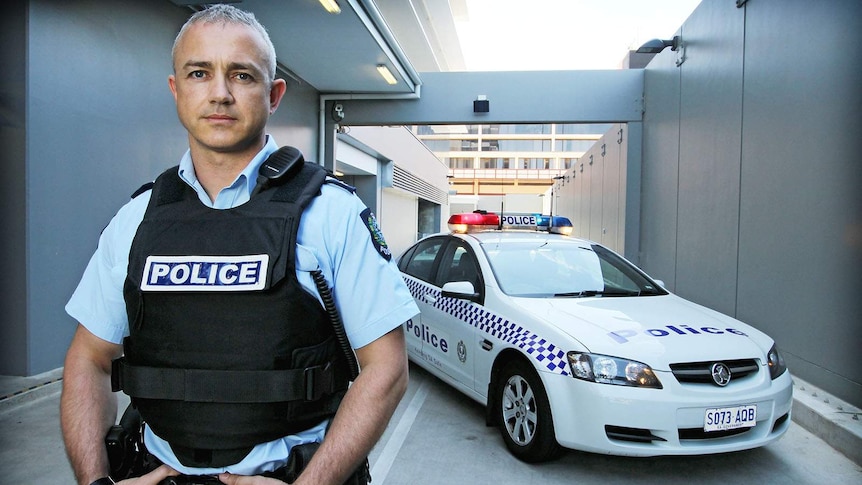 Eric Walsh stand in front of a police car wearing a police vest and uniform