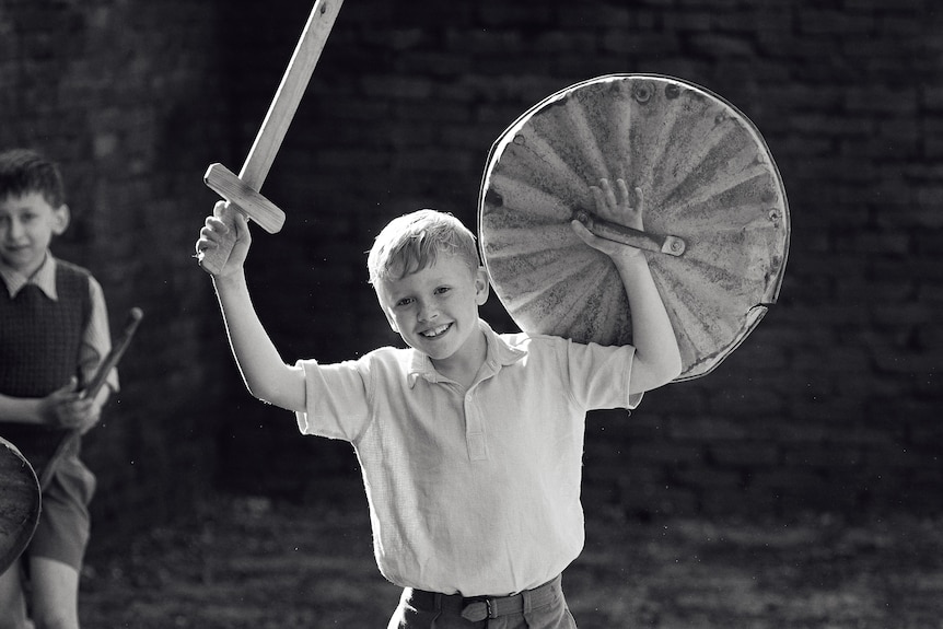 A black and white image of a boy in 60s-style clothing grinning as he holds up a toy sword and shield