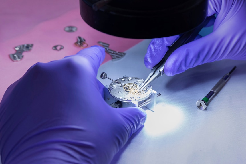 A close up image of a mechanical wristwatch being worked on by someone wearing purple gloves under a light