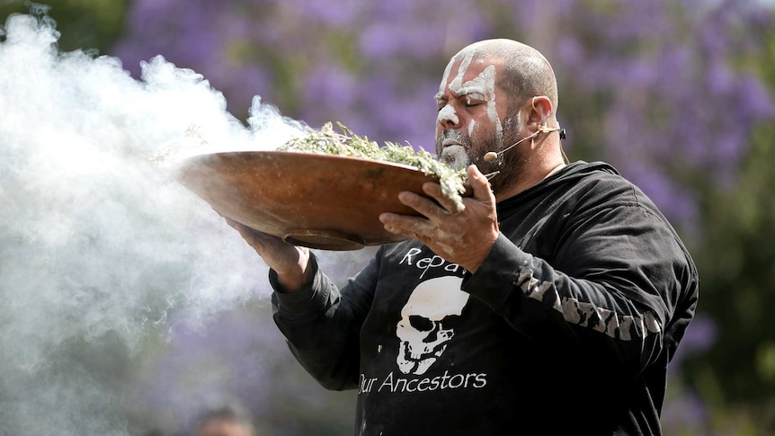 An Aboriginal man with his face painted holds aloft a large dish filled with smoking leaves with his eyes closed.