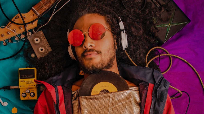 A man with sunglasses lies on a bed wearing headphones