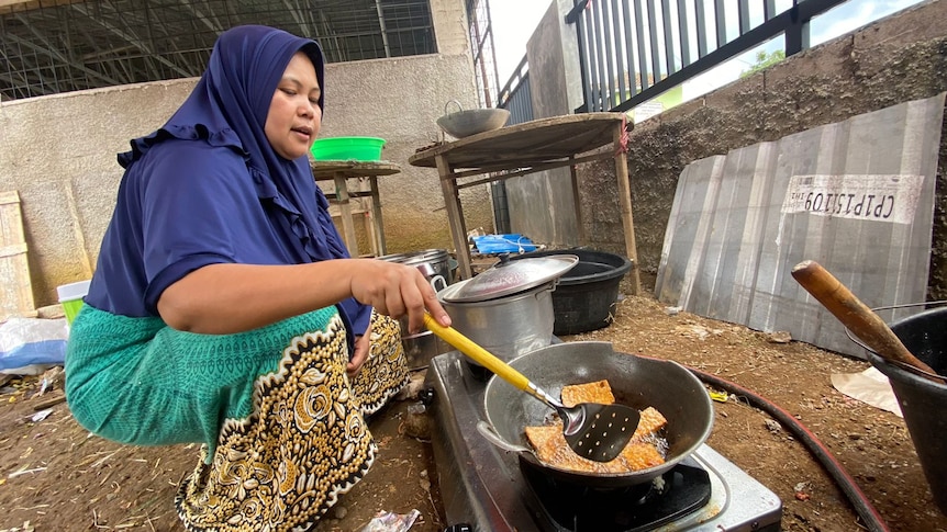 A woman wearing a blue hijab, sitting facing a frying pan, is cooking at the shelter.
