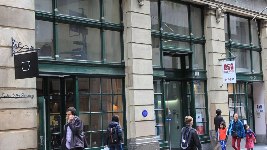 People walk past the ground level of Ross House, which is made from light grey stone with huge square windows with green frames.