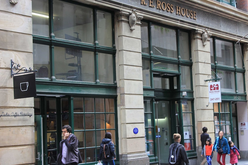 People walk past the ground level of Ross House, which is made from light grey stone with huge square windows with green frames.