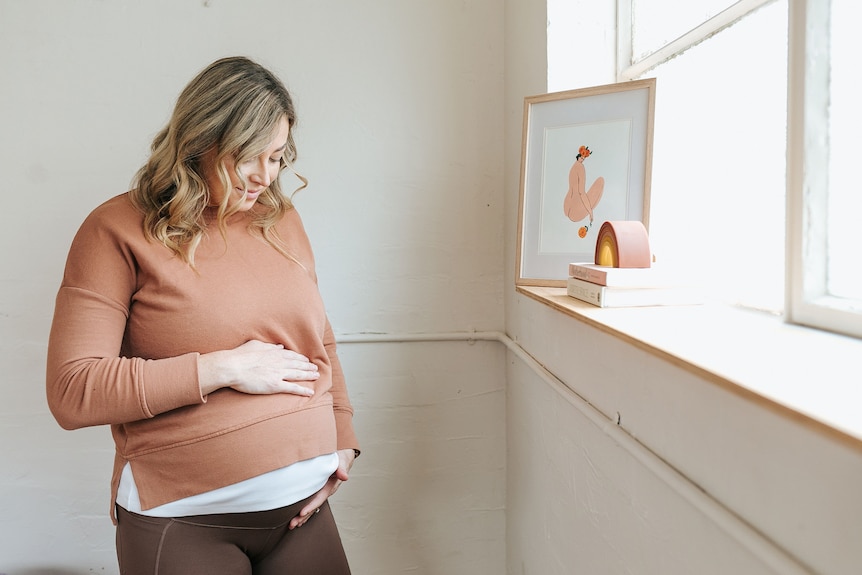 A heavily pregnant woman wearing a brown top looks at her belly