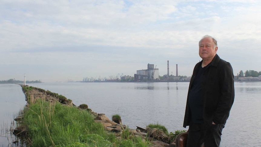 An older man stands on on a rocky pier in front of a wharf