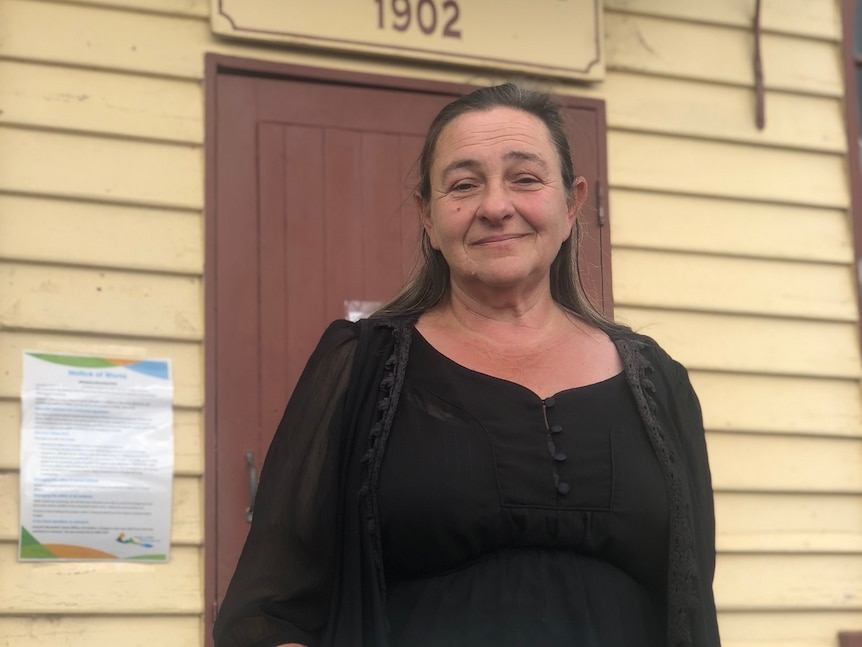 A late middle-aged woman stands outside a historic-looking building with a resigned look on her face.