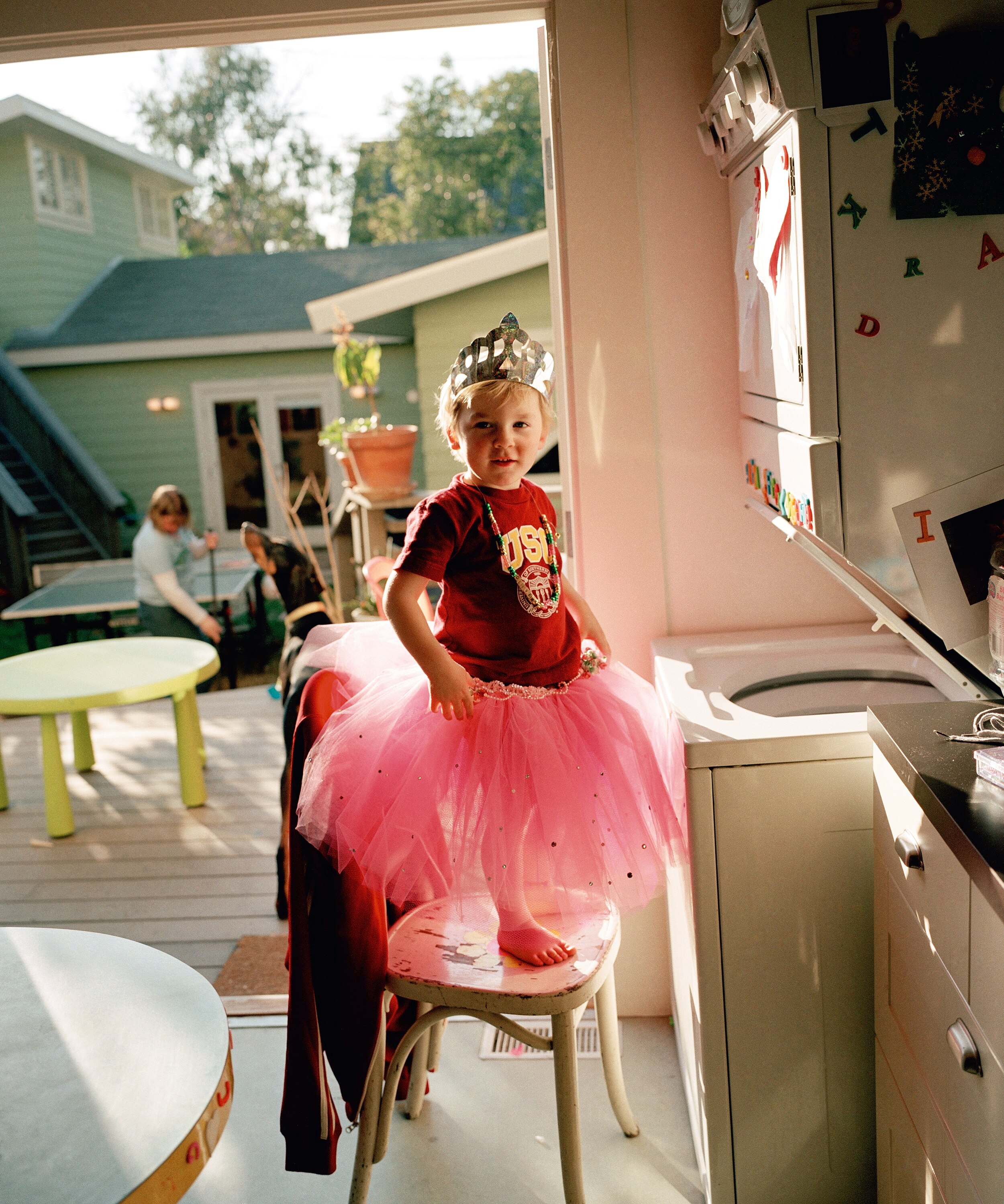 A blond-haired toddler wearing a t-shirt, tutu and tiara stands on a chair in front of a washing machine