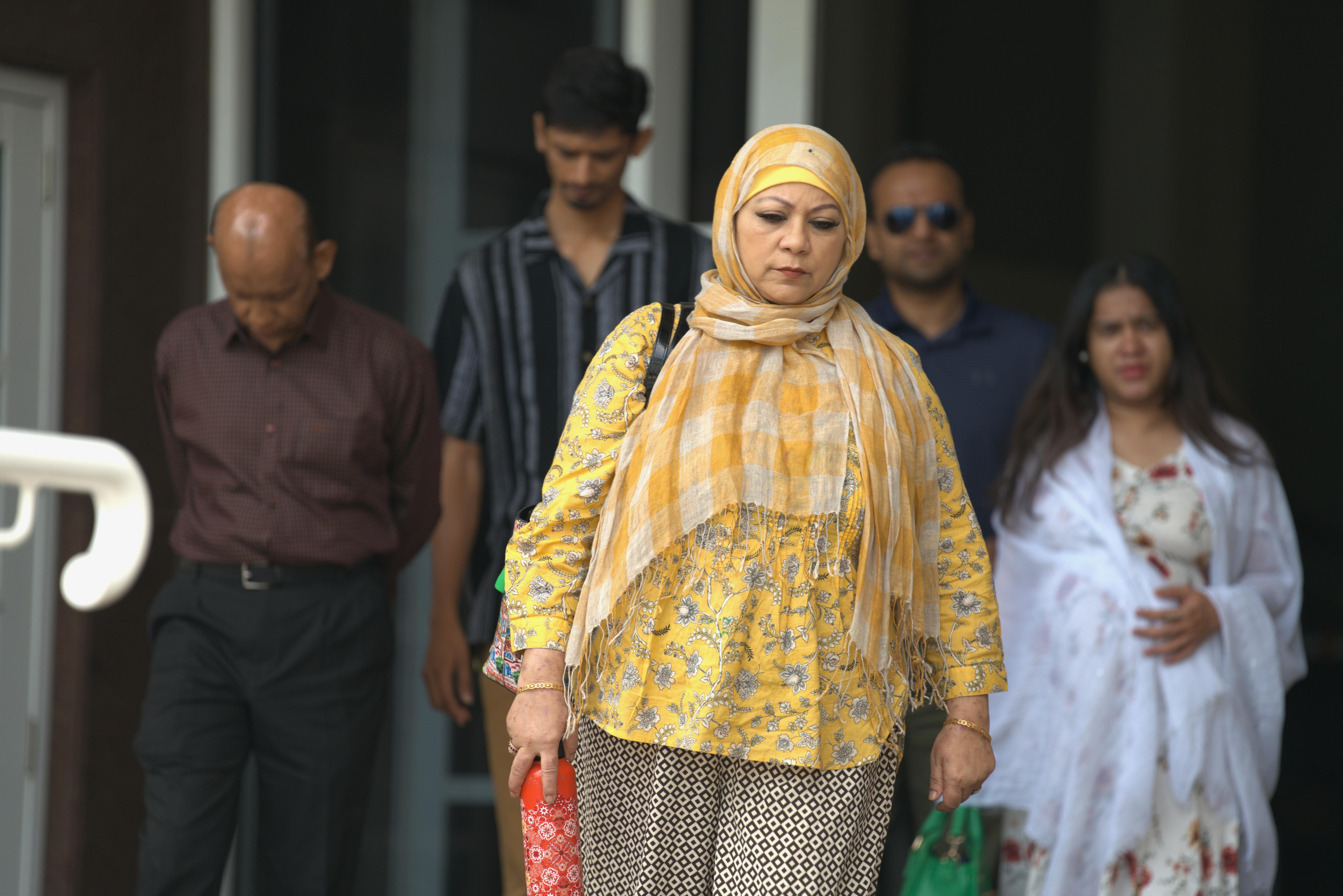 Una mujer con una blusa amarilla y un hijab caminando frente a la sala del tribunal con un grupo de personas detrás de ella.