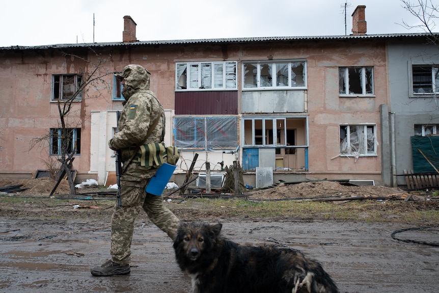 A man in full camo gear walks past a damaged apartment building, tailed by a black dog