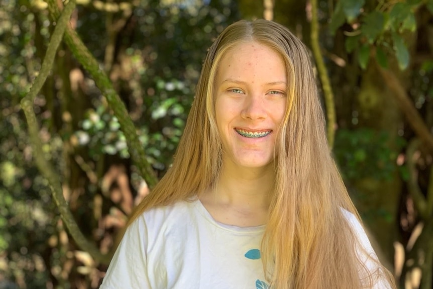 A young woman with blond hair and braces stands in front of green trees, smiling.