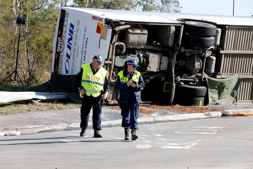 police crash investigators inspect the  road at the site of a crash. An overturned bus is in the background.