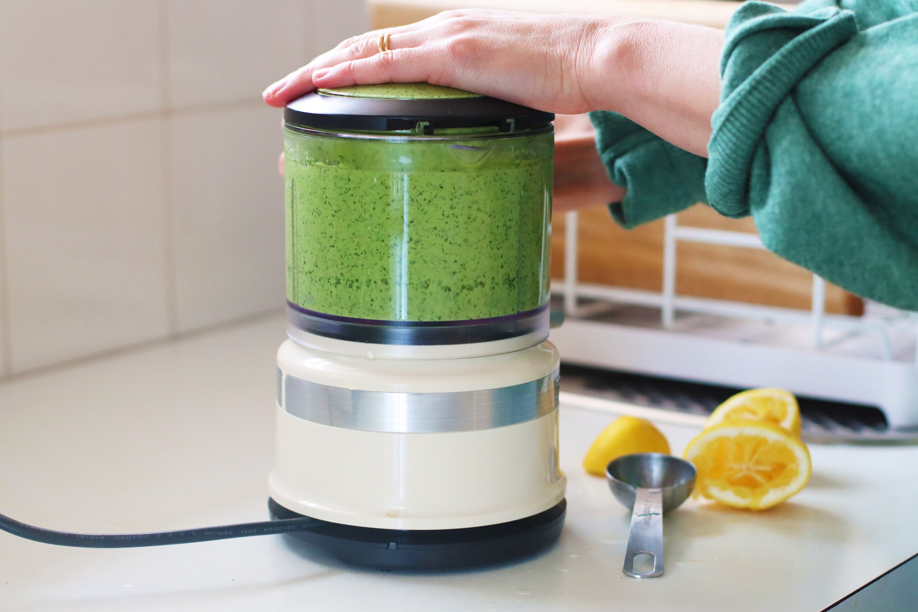 Close-up of hands on a small food processor blitzing up green tahini sauce. Cut lemons and the kitchen sink in background.