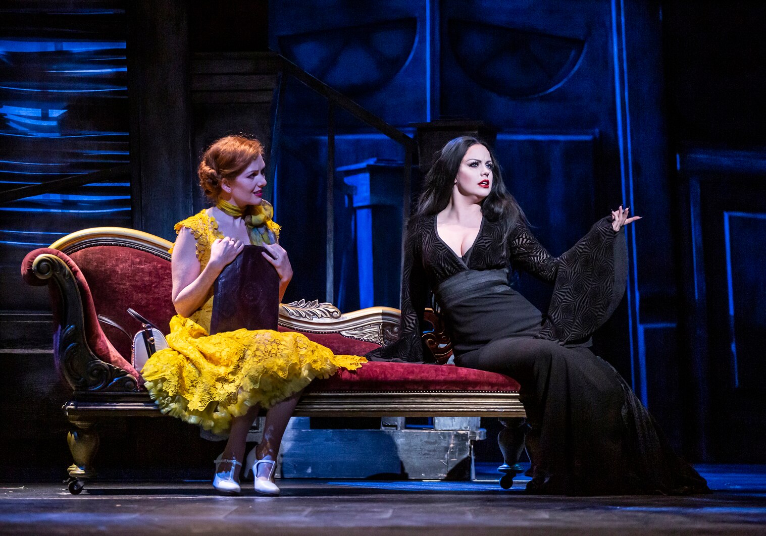 Two women on stage, one in bright yellow with red hari and the other pale with long black hair, wearing black. 