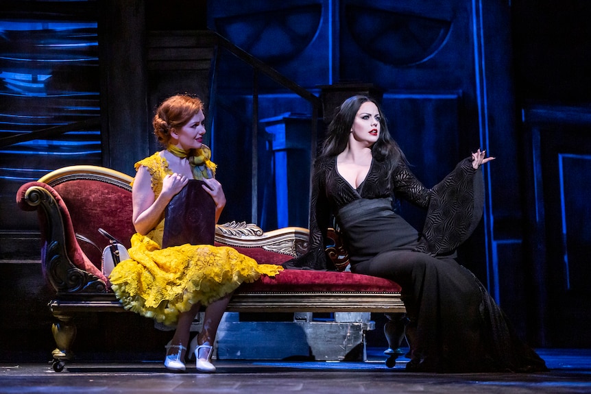 Two women on stage, one in bright yellow with red hari and the other pale with long black hair, wearing black. 