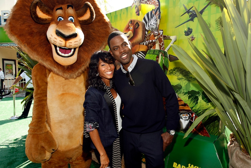 Chris Rock and Jada Pinkett Smith smile and embrace in front of a promotional poster for Madagascar: Escape 2 Africa
